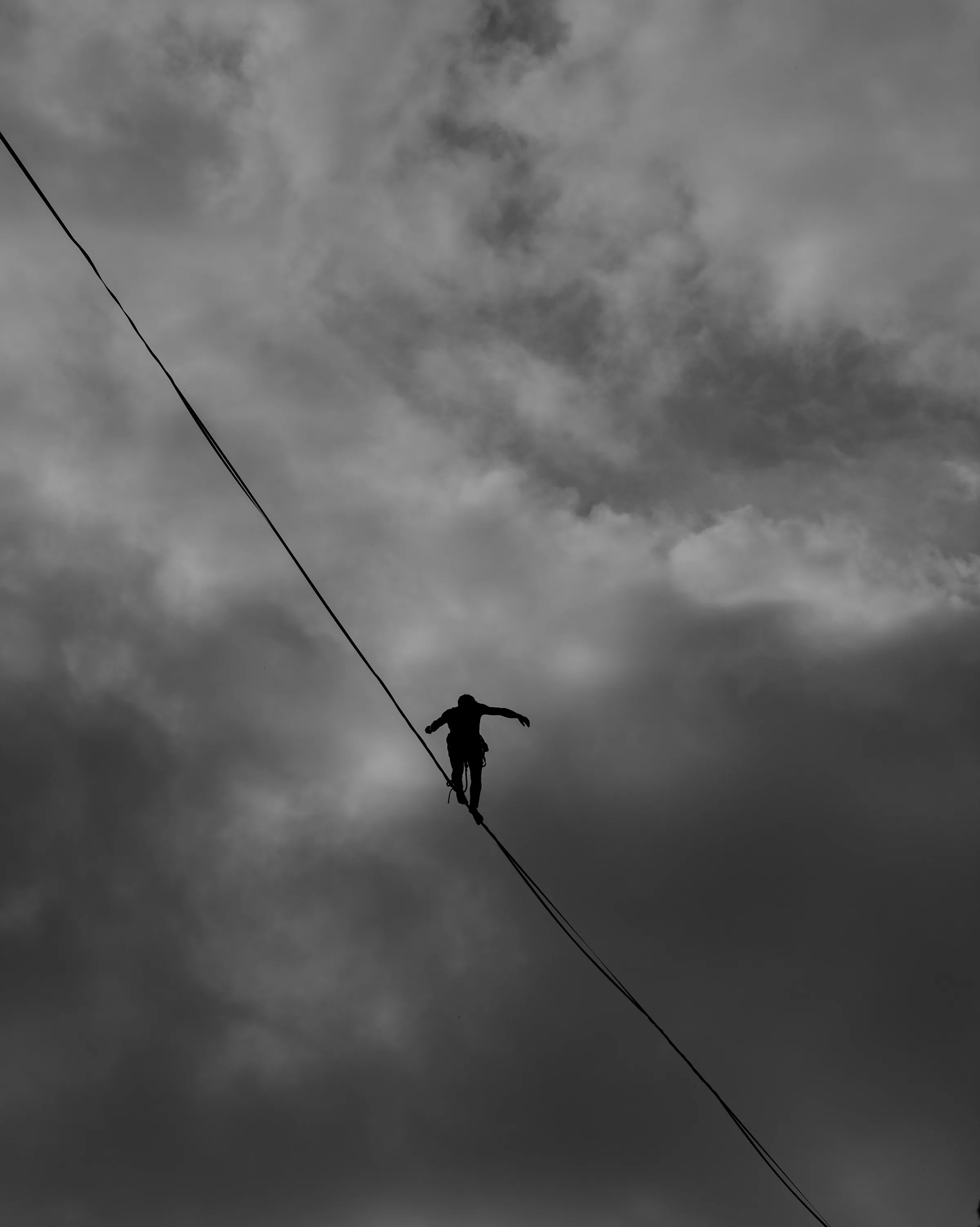 A lone figure balancing partway along a highline, high against a bank of grey cloud